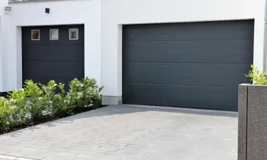 Dwie zamknięte szare bramy garażowe w białym budynku, małe okienka po lewej, podjazd i żywopłot