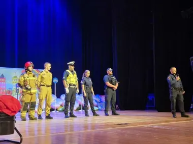 Funkcjonariusze służb mundurowych, w tym strażak, policjanci i strażnik miejski, stoją na scenie podczas wydarzenia edukacyjnego dla dzieci.
